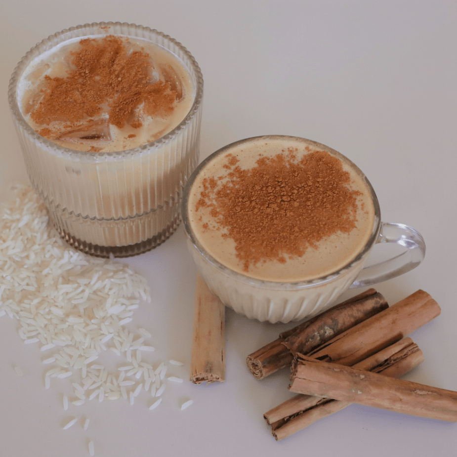 Two horchata drinks showing an iced horchata in a clear glass, and a hor horchata in a clear glass mug next to cinnamon sticks and rice pieces on the table.