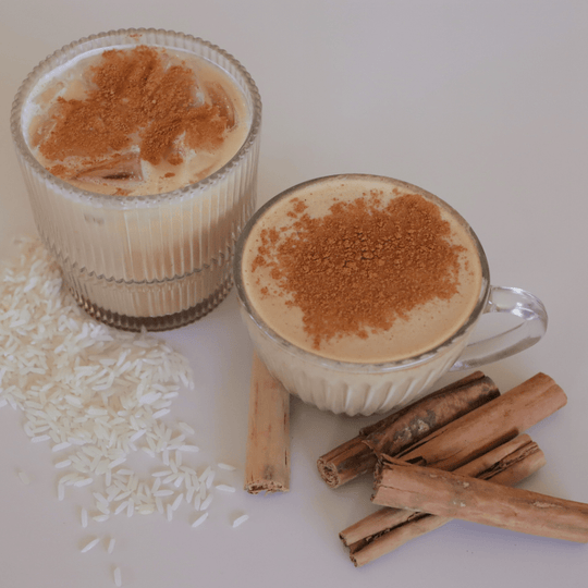 Two horchata drinks showing an iced horchata in a clear glass, and a hor horchata in a clear glass mug next to cinnamon sticks and rice pieces on the table.