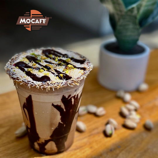 Dubai Chocolate Iced Coffee on table with Pistachios by plant with Mocafe logo