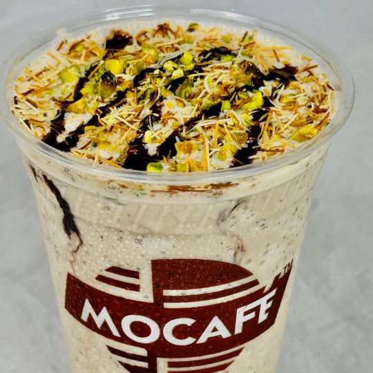 Close-up showing a MOCAFE Dubai Chocolate frappe showing garnish of chocolate sauce, kataifi, and crushed pistachio pieces. Chocolate chip pieces show in the base of the drink.