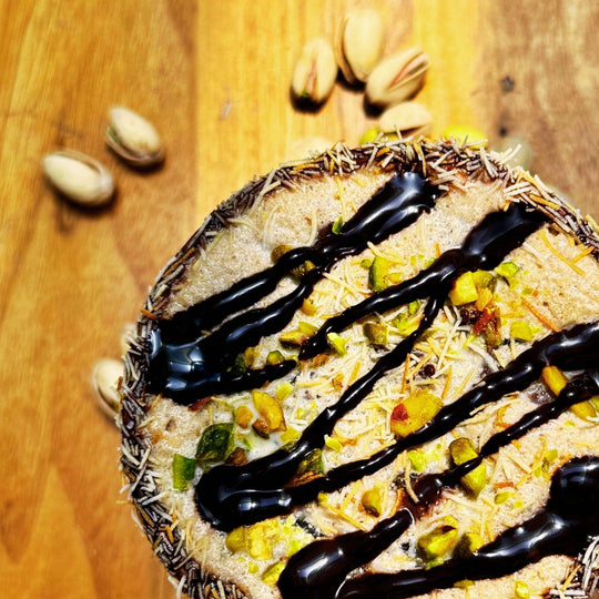 Dubai Chocolate Frappe drink top down view showing chocolate and kataifi rim, pistachio pieces and chocolate drizzle on top, and shelled pistachios on wooden table.