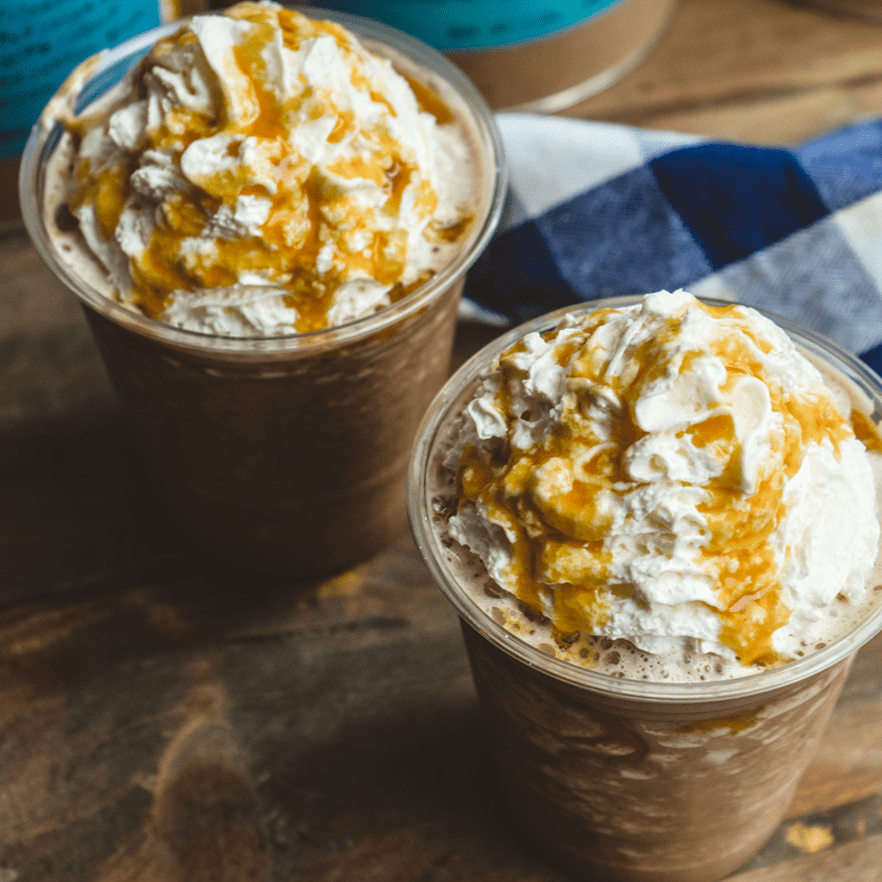 Two caramel frappes blended with ice with whipped cream on top and caramel sauce drizzle