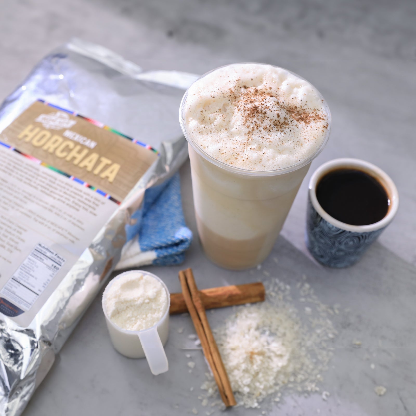 Horchata drink mix 3lb bag and scoop of white powder next to a made 16 oz Horchata frappe with a shot of espresso next to it and cinnamon sticks.