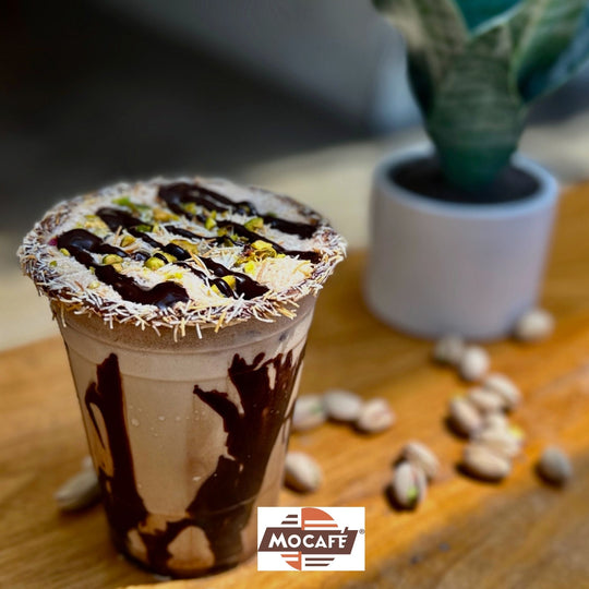 Mocafe Iced Dubai Chocolate Beverage with chocolate sauce inside cup shwoing pistachio and kataifi toppings pistachios and plant on table